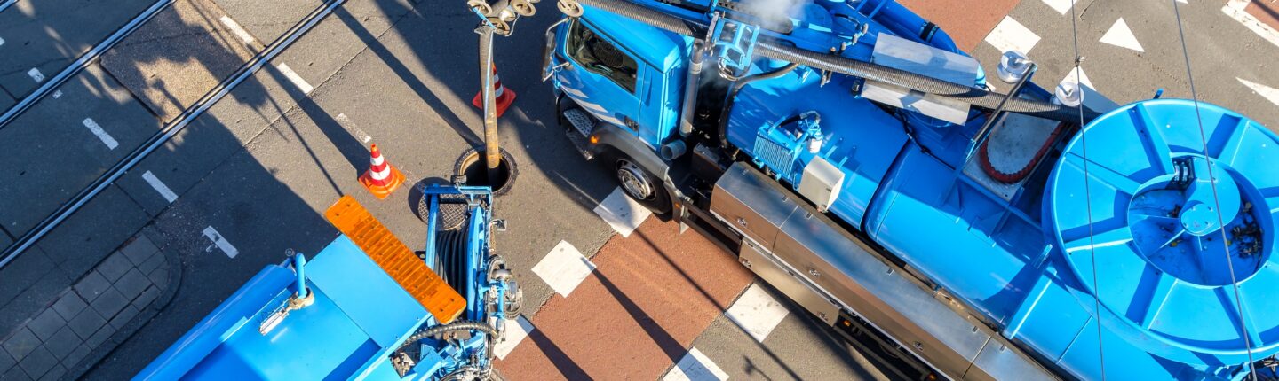 Twee wagens legen het riool op een kruispunt in Amsterdam; een voorbeeld van de uitvoering van de gemeentelijke rioolwatertaken